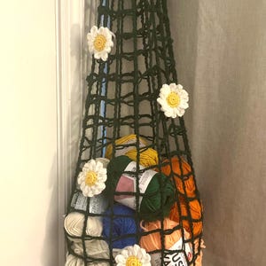 May include: A dark green, crocheted hanging basket filled with colorful yarn skeins. The basket is decorated with white and yellow daisy appliqués. The basket is suspended from a door frame.