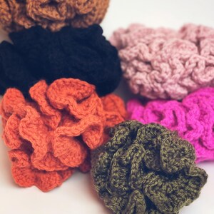 Könnte beinhalten: Eine Sammlung von sechs gehäkelten Haargummis in verschiedenen Farben, darunter Braun, Schwarz, Hellrosa, leuchtendes Orange, Pink und Olivgrün. Jedes Haargummi weist ein gerüschtes, strukturiertes Garnmuster mit einer runden, elastischen Form auf.