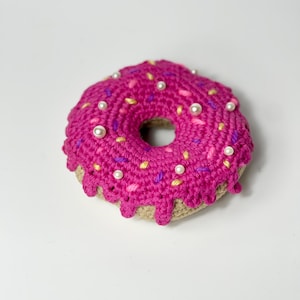 May include: A pink crocheted donut with white pearl sprinkles and yellow and purple sprinkles. The donut has a brown crocheted base.