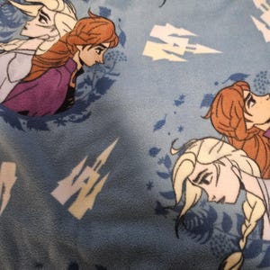 Puede incluir: Tela azul claro con un patr&oacute;n repetido de Elsa y Anna de la pel&iacute;cula Frozen. El dise&ntilde;o incluye im&aacute;genes de un castillo y copos de nieve. Los personajes tienen cabello rubio y casta&ntilde;o rojizo, y visten ropa morada y azul.