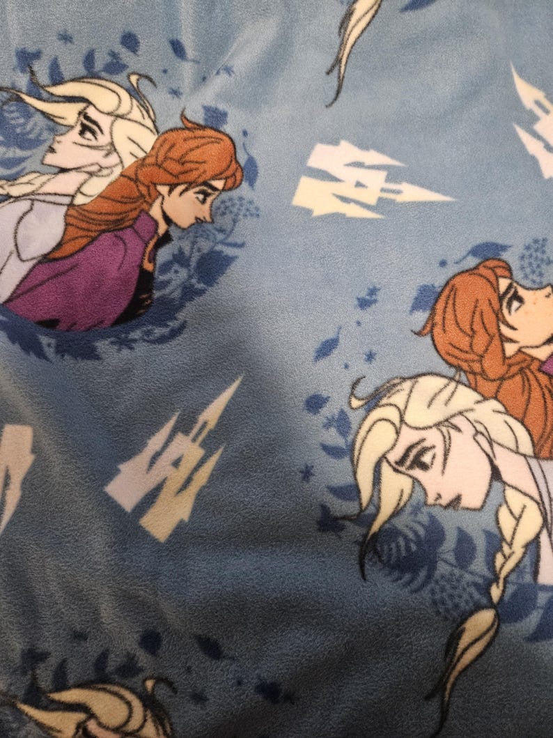 Puede incluir: Tela azul claro con un patr&oacute;n repetido de Elsa y Anna de la pel&iacute;cula Frozen. El dise&ntilde;o incluye im&aacute;genes de un castillo y copos de nieve. Los personajes tienen cabello rubio y casta&ntilde;o rojizo, y visten ropa morada y azul.