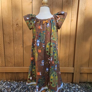 May include: A brown, short-sleeved dress with a whimsical forest print featuring foxes, pumpkins, and trees. The dress has a white lace trim at the hem and a small orange bow at the neckline. The dress is displayed on a mannequin.