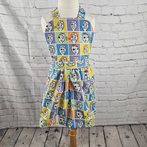 May include: A colorful child's apron with a repeating pattern of the character Forky from the Toy Story movie franchise. The apron has a halter neck style and a gathered skirt.