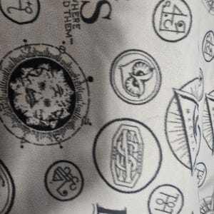 Puede incluir: Tela gris claro con un patr&oacute;n repetido de emblemas circulares y texto. Los emblemas incluyen una cabeza de le&oacute;n, un b&uacute;ho y dise&ntilde;os de br&uacute;jula. El texto incluye las palabras "FANTASTIC BEASTS" y "AND WHERE TO FIND THEM."