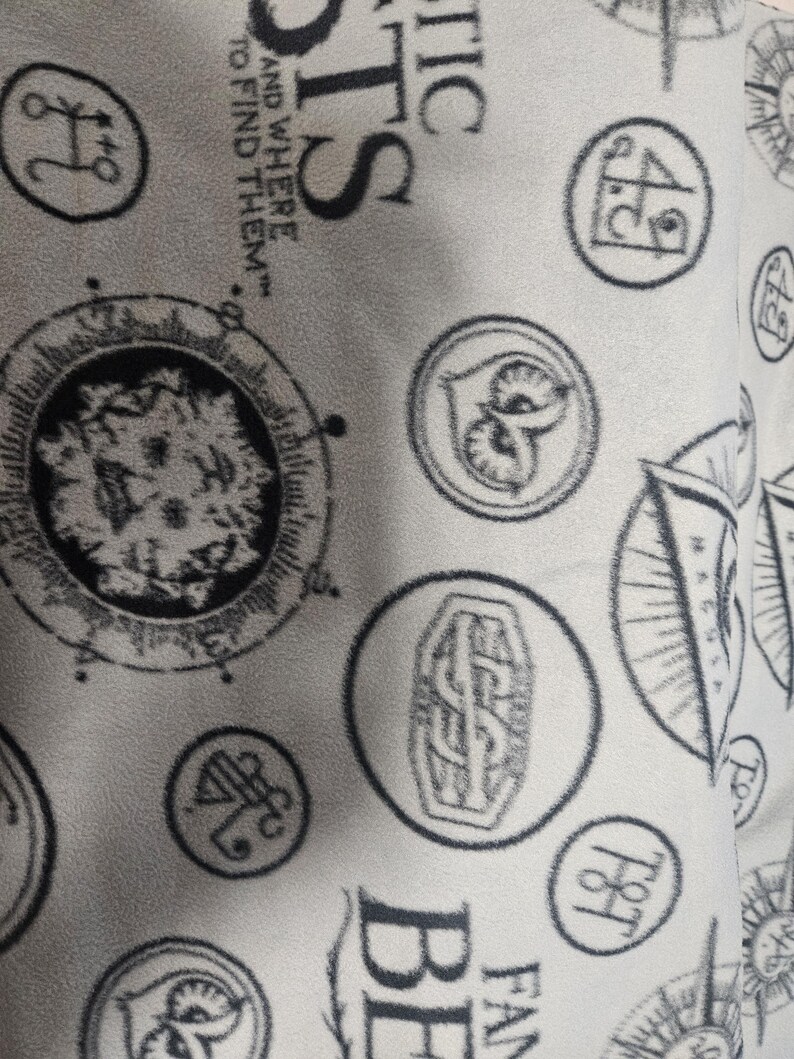 Puede incluir: Tela gris claro con un patr&oacute;n repetido de emblemas circulares y texto. Los emblemas incluyen una cabeza de le&oacute;n, un b&uacute;ho y dise&ntilde;os de br&uacute;jula. El texto incluye las palabras "FANTASTIC BEASTS" y "AND WHERE TO FIND THEM."