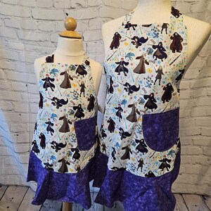 May include: Two aprons, one adult and one child-sized, displayed on mannequins. The aprons feature a white background with a repeating pattern of figures in black, brown, and gray. Each apron has a purple pocket and a purple ruffled hem.