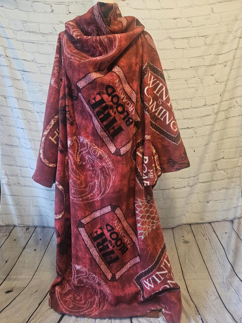 Puede incluir: Una manta con capucha de color burdeos con un patr&oacute;n repetido del sigilo Targaryen y las palabras "Fire and Blood". La manta tambi&eacute;n presenta la frase "Winter is Coming".