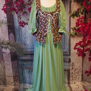 May include: A green dress with a black and orange patterned bodice and long sleeves. The bodice has a square neckline and white lacing. The dress is displayed on a mannequin. The background features a rustic door and red flowers.