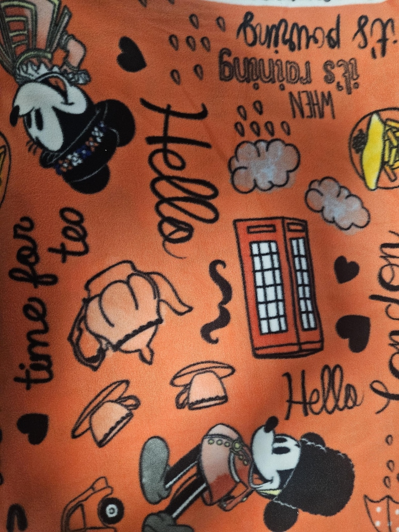 Puede incluir: Tela naranja con ilustraciones de dibujos animados de Mickey Mouse, las palabras "Hello", "time for tea" y "it's raining". El dise&ntilde;o incluye corazones, nubes y una cabina telef&oacute;nica roja. Un dise&ntilde;o divertido y juguet&oacute;n.