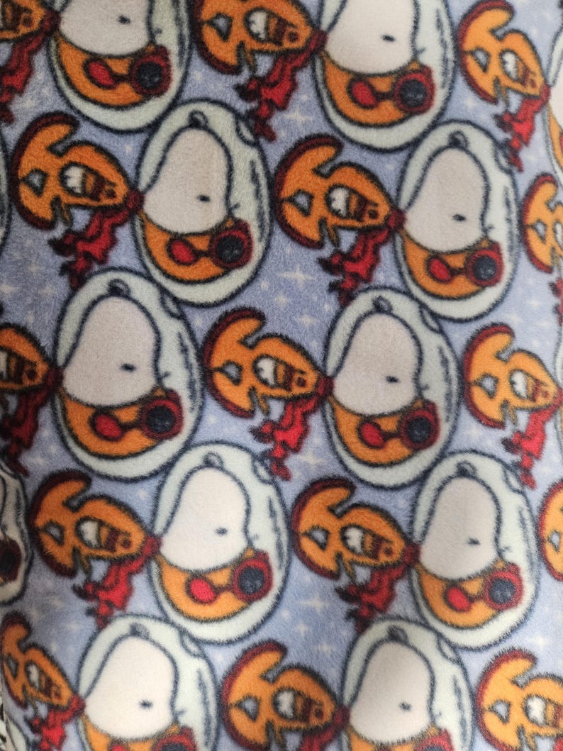 Puede incluir: Tejido polar azul claro con un patr&oacute;n repetido de personajes de dibujos animados. Cada personaje est&aacute; dentro de un &oacute;valo blanco con detalles en naranja y rojo. El fondo tiene un sutil patr&oacute;n de estrellas.