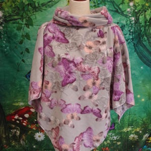 May include: A gray poncho with a cowl neck, adorned with a pattern of purple and pink butterflies and flowers. The poncho is displayed on a mannequin against a backdrop of a whimsical forest scene.