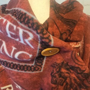 Puede incluir: Un chaleco de forro polar rojo y negro con un cierre de bot&oacute;n de madera grande. El chaleco presenta un patr&oacute;n repetitivo con texto blanco y dise&ntilde;os gr&aacute;ficos negros. El chaleco est&aacute; drapeado sobre un maniqu&iacute;.