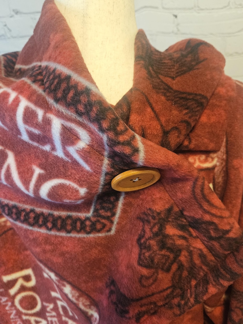 Puede incluir: Un chaleco de forro polar rojo y negro con un cierre de bot&oacute;n de madera grande. El chaleco presenta un patr&oacute;n repetitivo con texto blanco y dise&ntilde;os gr&aacute;ficos negros. El chaleco est&aacute; drapeado sobre un maniqu&iacute;.