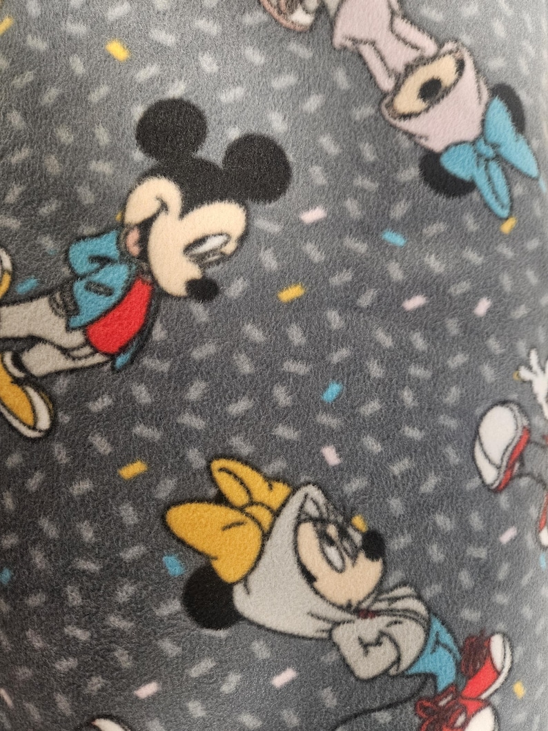 Puede incluir: Tejido polar gris con un patr&oacute;n repetido de Mickey y Minnie Mouse. Los personajes se representan en varias poses, vistiendo sus trajes ic&oacute;nicos, con formas de confeti dispersas en el fondo.