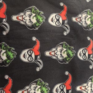 Puede incluir: Tela negra con un patr&oacute;n repetido de las caras de Joker y Harley Quinn. La cara del Joker tiene pelo verde y una amplia sonrisa. La cara de Harley Quinn tiene un sombrero de buf&oacute;n rojo y negro y maquillaje blanco.