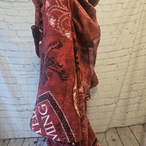 Puede incluir: Una capa-manta con capucha de color burdeos con un dise&ntilde;o estampado. La capa tiene una capucha y un bolsillo delantero. La tela tiene una textura suave y afelpada. El dise&ntilde;o incluye adornos y texto.