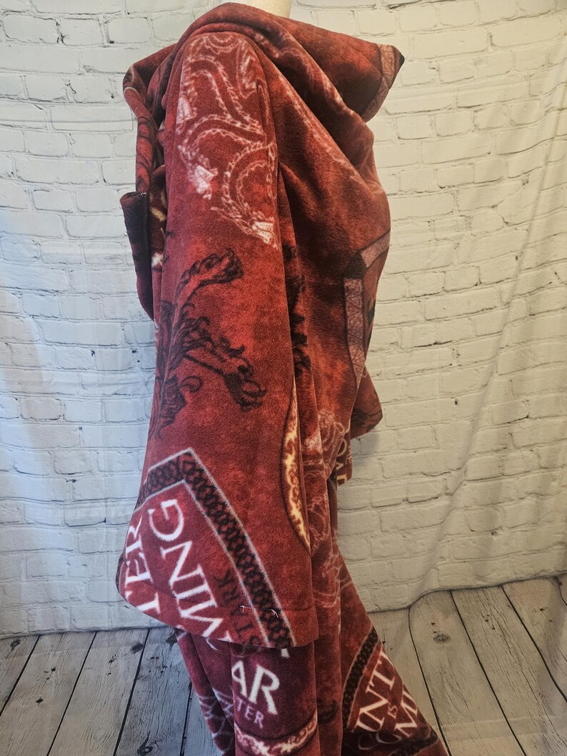 Puede incluir: Una capa-manta con capucha de color burdeos con un dise&ntilde;o estampado. La capa tiene una capucha y un bolsillo delantero. La tela tiene una textura suave y afelpada. El dise&ntilde;o incluye adornos y texto.