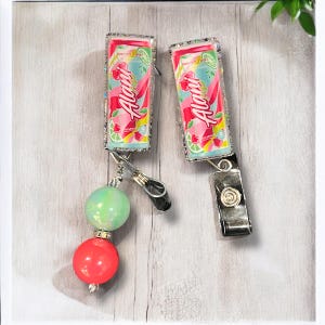 May include: Two pink and blue Aloha-branded beverage can-shaped badge holders with glitter accents. Each holder has a silver clip and a beaded chain with a green and a red bead.