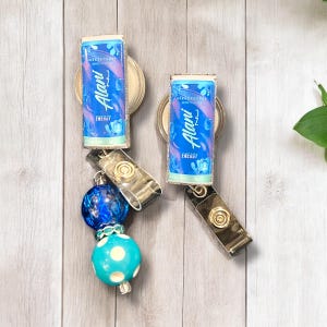 May include: Two blue and silver retractable badge holders with a blue and white polka dot bead and a blue glass bead. The badge holders feature a printed image of an energy drink can with the text "Alani" and "Energy".