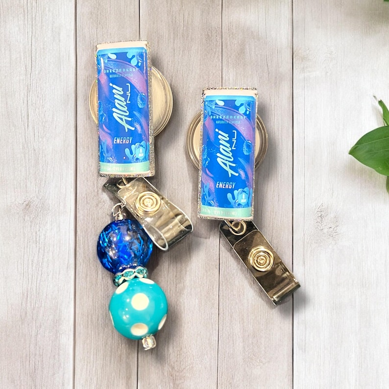 May include: Two blue and silver retractable badge holders with a blue and white polka dot bead and a blue glass bead. The badge holders feature a printed image of an energy drink can with the text "Alani" and "Energy".