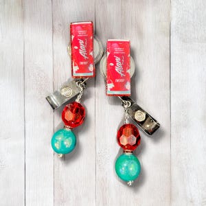 May include: Two red and white Alani Nu energy drink can badge reels with turquoise and red beads.