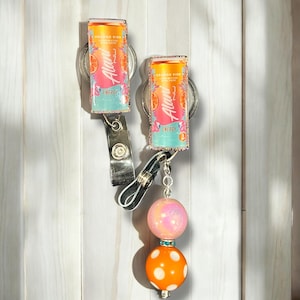 May include: Two colourful keychains featuring a can of  "Auii" Orange Kiwi Energy drink. The keychains are attached to a pink and white swirl bead and an orange bead with white polka dots.