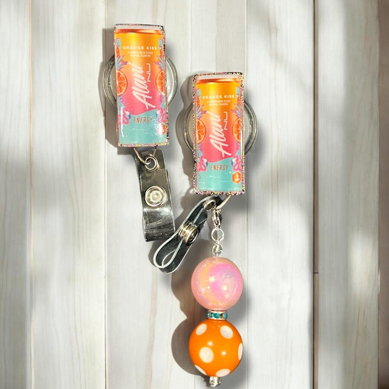 May include: Two colourful keychains featuring a can of  "Auii" Orange Kiwi Energy drink. The keychains are attached to a pink and white swirl bead and an orange bead with white polka dots.