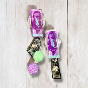 May include: Two purple and green retractable badge reels with a green and purple iridescent finish. The reels feature a graphic of a can of Alani Nu energy drink with the text "Cosmic Stardust" and "Alani Nu" printed on the can. The reels are attached to a silver chain with a green and a purple iridescent bead.