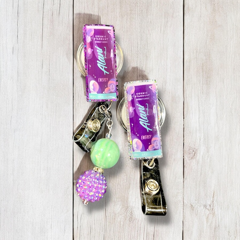 May include: Two purple and green retractable badge reels with a green and purple iridescent finish. The reels feature a graphic of a can of Alani Nu energy drink with the text "Cosmic Stardust" and "Alani Nu" printed on the can. The reels are attached to a silver chain with a green and a purple iridescent bead.