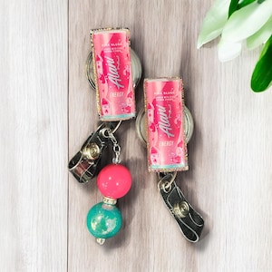 May include: Two pink and gold sparkly keychains with a pink and white can design. The can says "Alani" and "Pink Blush" with a pink and white design. The keychains have a black clip and a pink and turquoise bead.