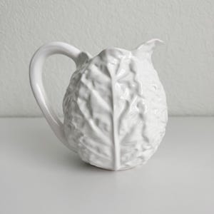 May include: A white ceramic pitcher featuring a textured surface that mimics the appearance of overlapping cabbage leaves. The vessel has a curved handle on the side and a shaped pouring spout at the top, finished with a glossy glaze.
