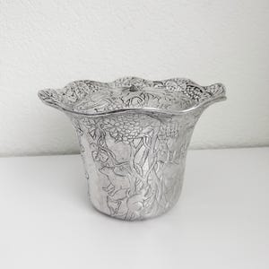 May include: A silver-toned metal bowl with a scalloped rim and a matching lid. The exterior surface features an embossed relief design depicting grapevines, clusters of grapes, and small animals including foxes among the foliage.