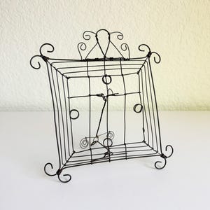 May include: A dark brown wire display stand featuring a square frame with a grid pattern. The structure includes decorative curled wire accents at each corner and a central support mechanism with a small hook for holding items upright.