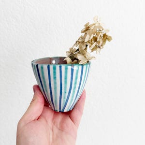 May include: A small, conical ceramic wall planter featuring vertical stripes in alternating shades of teal and dark blue. The vessel has a single circular hole near the rim for mounting and contains a dried, light brown hydrangea flower stem.