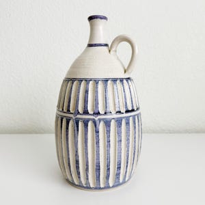 May include: A ceramic jug with a narrow neck and a single handle. The body features a cream-colored base with vertical, recessed grooves painted in a deep blue glaze. A horizontal blue band separates the upper and lower sections of the vessel.