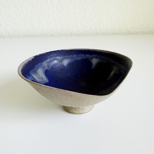 May include: A ceramic bowl with a wide, irregular rim and a pedestal base. The interior features a deep, glossy cobalt blue glaze that transitions into a lighter, mottled grey-white tone near the edges. The exterior has a textured, matte beige finish.