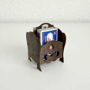 May include: A dark brown, hammered metal matchbox holder with decorative cutouts and a scalloped top edge. Inside the holder sits a rectangular matchbox with blue and white branding that reads 32 STRIKE ANYWHERE MATCHES and KEEP AWAY FROM CHILDREN.