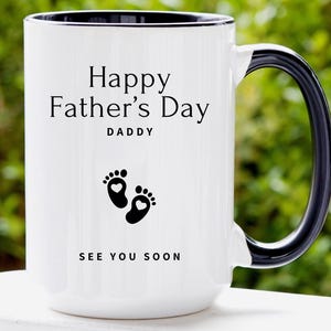 May include: White ceramic mug with a black handle and rim. The mug features the text "Happy Father's Day DADDY" and the words "SEE YOU SOON" below a pair of black baby footprints with heart-shaped details.