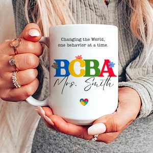 May include: White ceramic coffee mug with a black and white design. The mug reads "Changing the World, one behavior at a time." and "BCBA Mrs. Smith". The letters in "BCBA" are in different colors and have puzzle pieces on them. There is a small blue and yellow puzzle piece heart at the bottom of the mug.