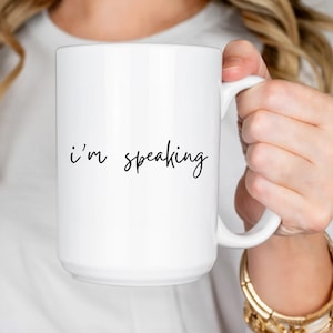 May include: White ceramic mug with the phrase "I'm speaking" in black script. The mug has a large handle and is held by a person. The mug is a simple, yet bold statement piece.
