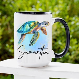 May include: White ceramic mug with a black rim and handle. The mug features a watercolor illustration of a sea turtle swimming in blue water with gold glitter accents. The name "Samantha" is written in black cursive below the turtle.