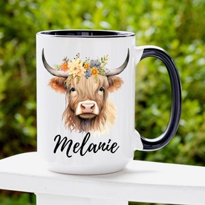 May include: White ceramic mug with a black rim and handle. The mug features a watercolor illustration of a Highland cow with a floral crown. The name "Melanie" is written in black cursive below the cow.