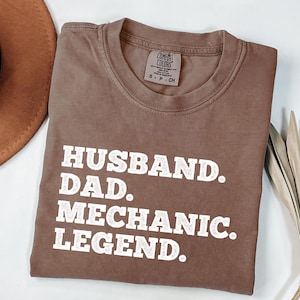 Puede incluir: Una camiseta marr&oacute;n con el texto "Husband. Dad. Mechanic. Legend." impreso en blanco.