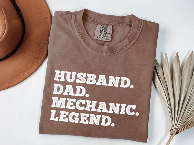 Puede incluir: Una camiseta marr&oacute;n con el texto "Husband. Dad. Mechanic. Legend." impreso en blanco.
