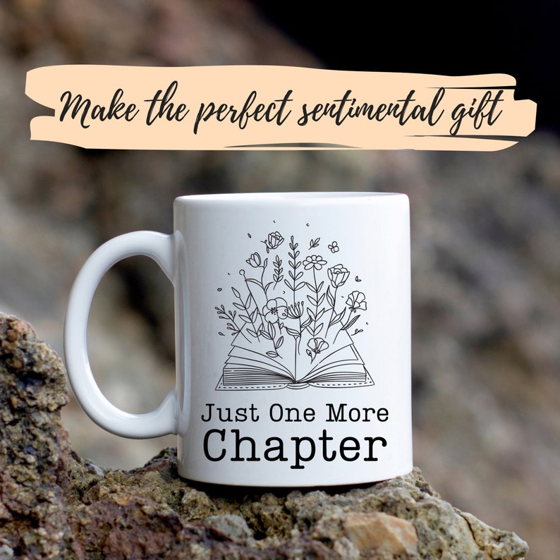Puede incluir: Taza de cer&aacute;mica blanca con un dibujo en l&iacute;nea negra de un libro abierto con flores que crecen de &eacute;l. El texto "Just One More Chapter" est&aacute; impreso debajo del libro.