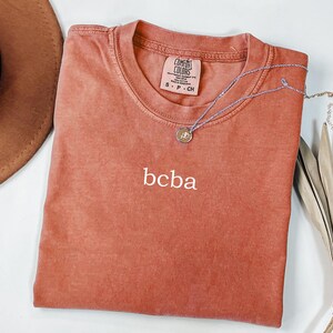 Op de afbeelding: Een roestkleurig T-shirt met de tekst "bcba" geborduurd op de voorkant. Het shirt is van Comfort Colors en is maat S-P-CH.