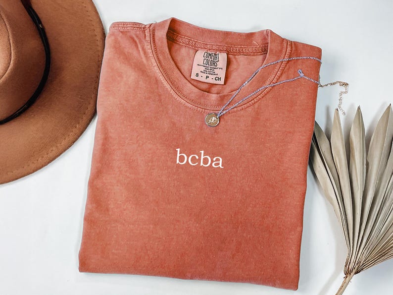 Op de afbeelding: Een roestkleurig T-shirt met de tekst "bcba" geborduurd op de voorkant. Het shirt is van Comfort Colors en is maat S-P-CH.