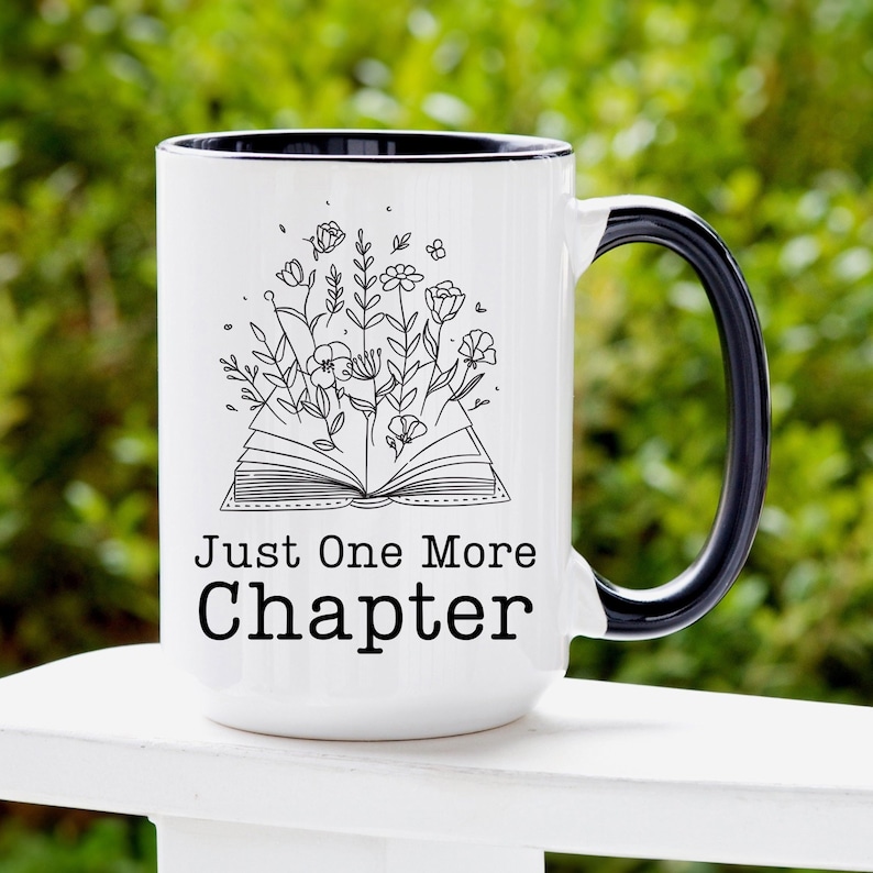 Puede incluir: Taza de caf&eacute; de cer&aacute;mica blanca con borde y asa negros. La taza presenta una ilustraci&oacute;n en blanco y negro de un libro abierto con flores que crecen de &eacute;l. El texto "Just One More Chapter" est&aacute; impreso en negro debajo de la ilustraci&oacute;n.
