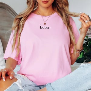 Op de afbeelding: Een oversized roze t-shirt met de tekst "bcba" erop gedrukt. Het shirt wordt gedragen door een persoon met lang blond haar.
