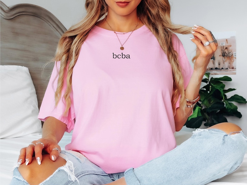 Op de afbeelding: Een oversized roze t-shirt met de tekst "bcba" erop gedrukt. Het shirt wordt gedragen door een persoon met lang blond haar.